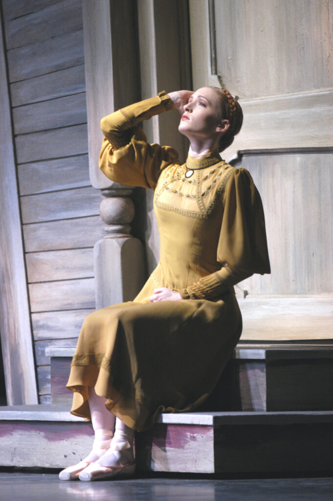 Murphy, wearing a long-sleeved yellow dress, sits on a wooden stoop onstage and looks up with a pensive look on her face. She runs her right hand along the side of her temple.