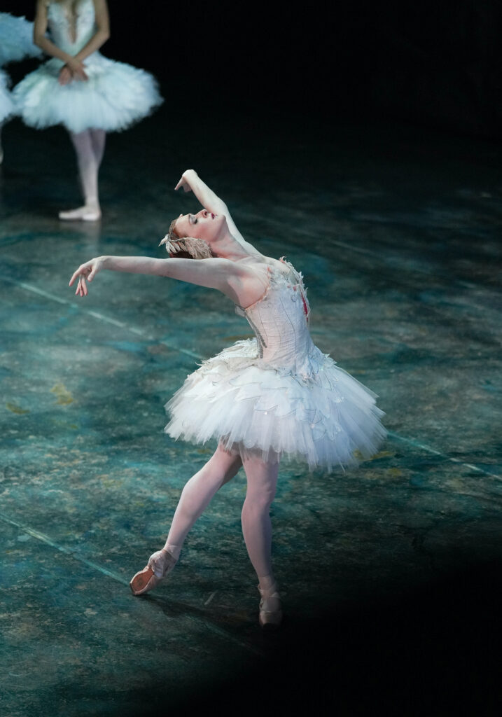 Murphy, wearing a white tutu with feathered embellishments, feathered headpiece, pink tights, and pink pointe shoes, stands in tendu derriere facing upstage. She opens her arms into a high V-shape and arches back, looking towards the ceiling.