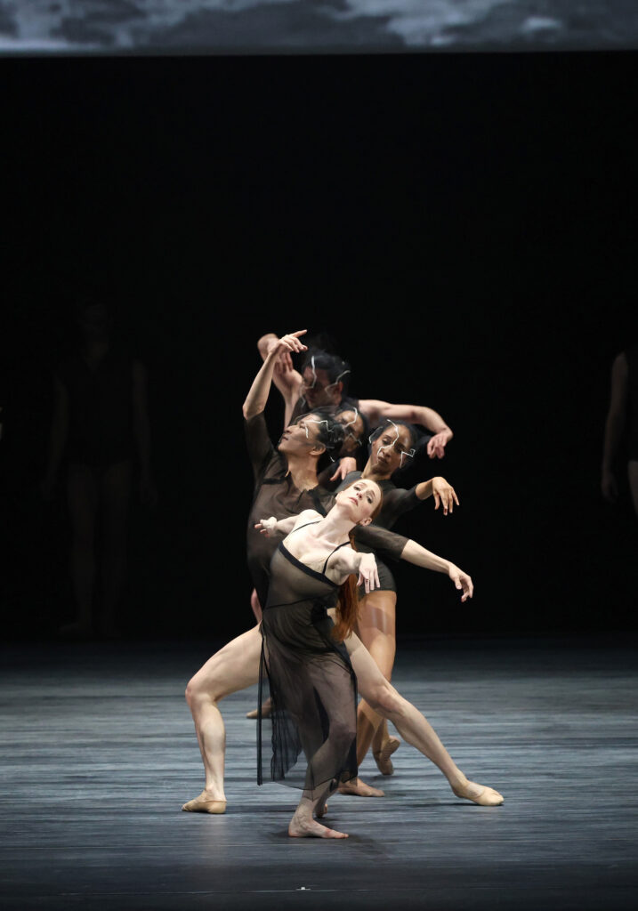 Gillian Murphy, in a mesh black dress, reaches out towards the audience with her arm outstretched. A small group of dancers in black biketards and mesh facemasks stand behind her in a formation.
